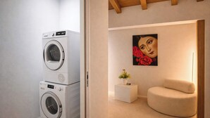 Een wasruimte met gestapelde wasmachines, een klein wit bijzettafeltje met een plant, en een beige fauteuil.