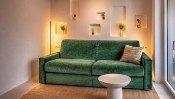 Un canapé vert, une petite table d’appoint blanche, une étagère murale ornée de vases et une fenêtre offrant une belle vue.