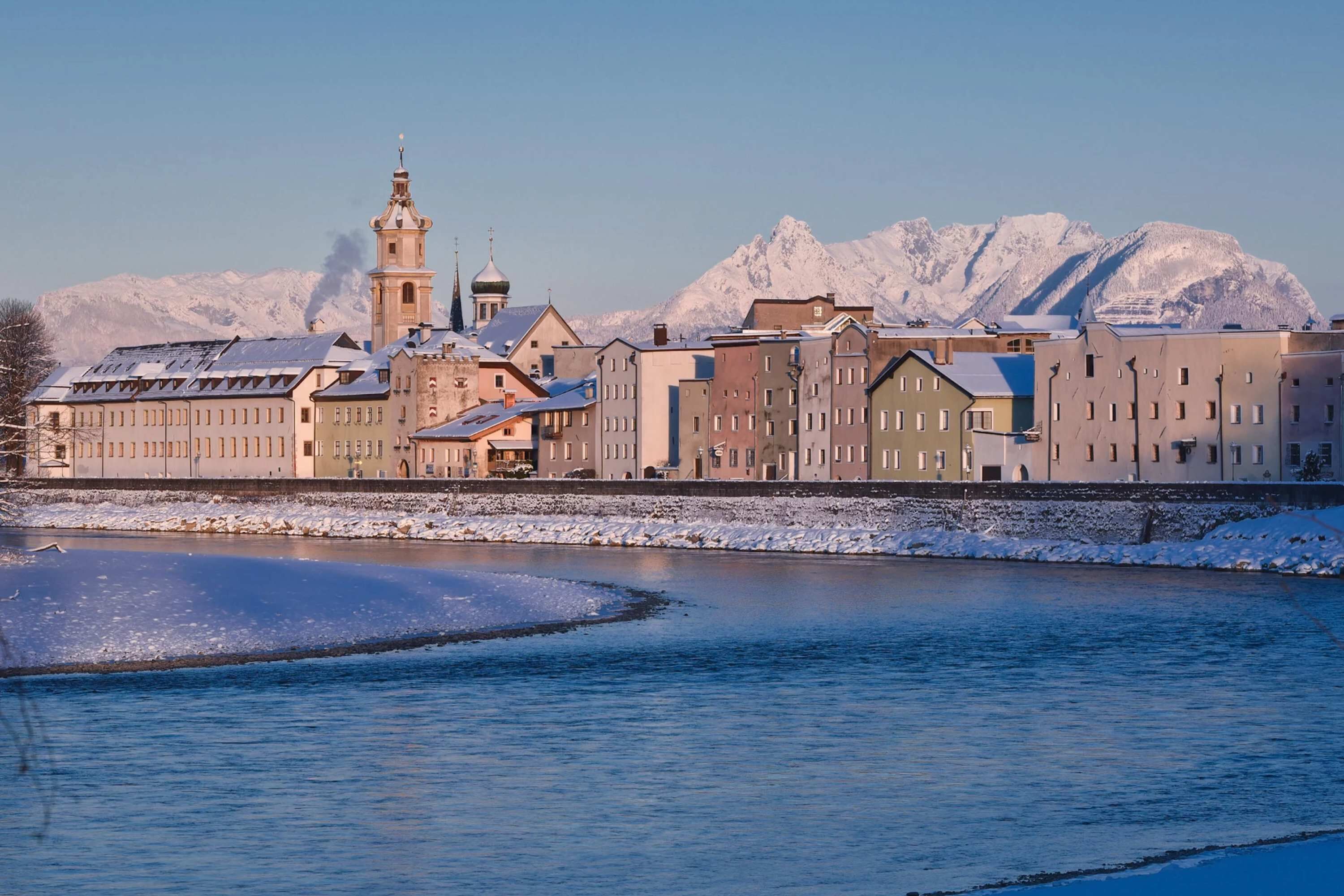 Eine Flussstadt mit historischen Gebäuden, einem markanten Kirchturm und schneebedeckten Bergen im Hintergrund.