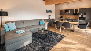 A modern living room with a grey sofa, a dining area with a table and chairs, and a kitchen with dark cabinets and appliances. - ZILLERHEART – Apartment with Balcony & Mountain View (Fügen)