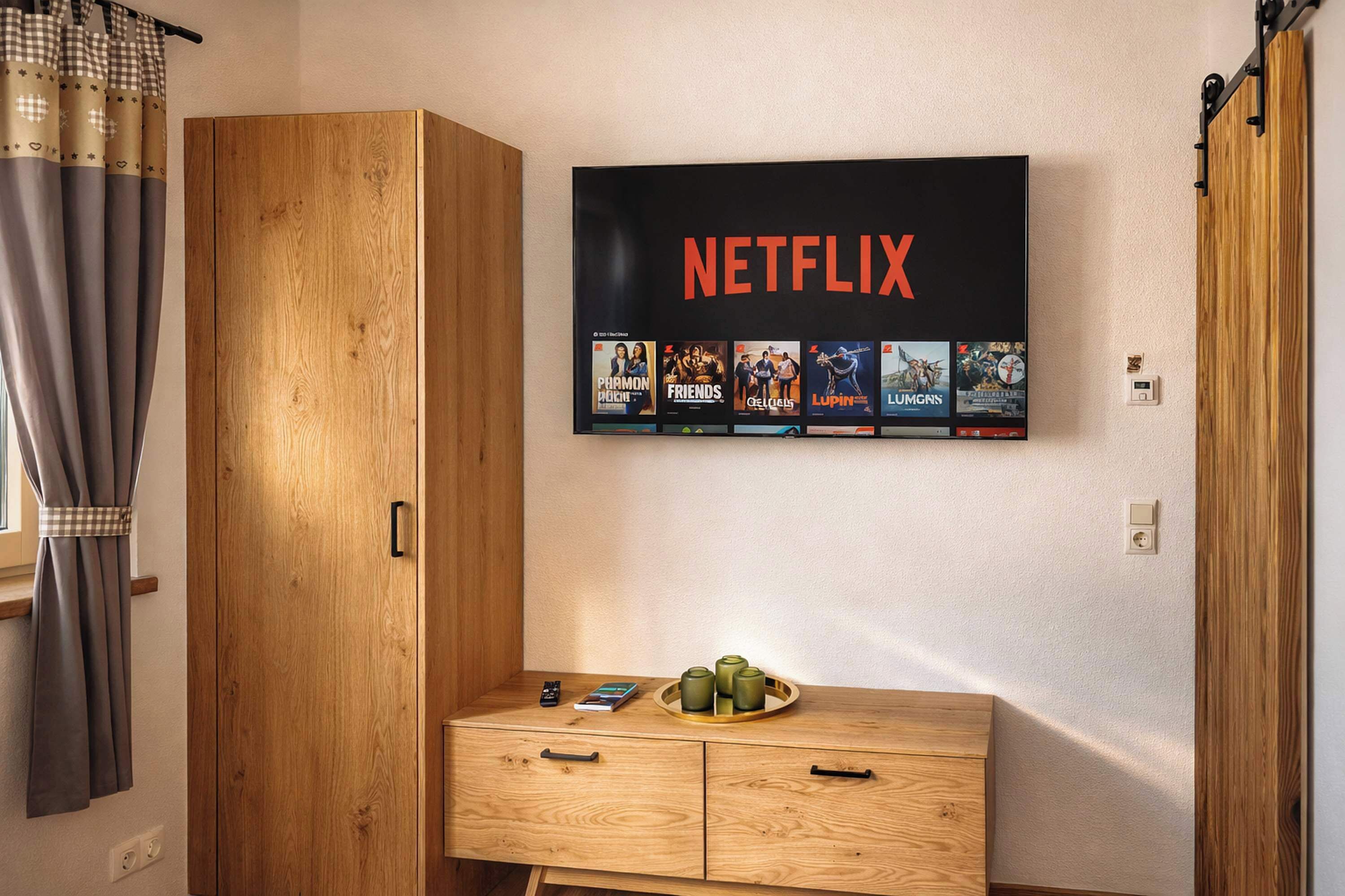 Ein Zimmer mit einem Holzschrank, einer Holzkommode und einem an der Wand montierten Netflix-Bildschirm, der verschiedene Sendungen zeigt.