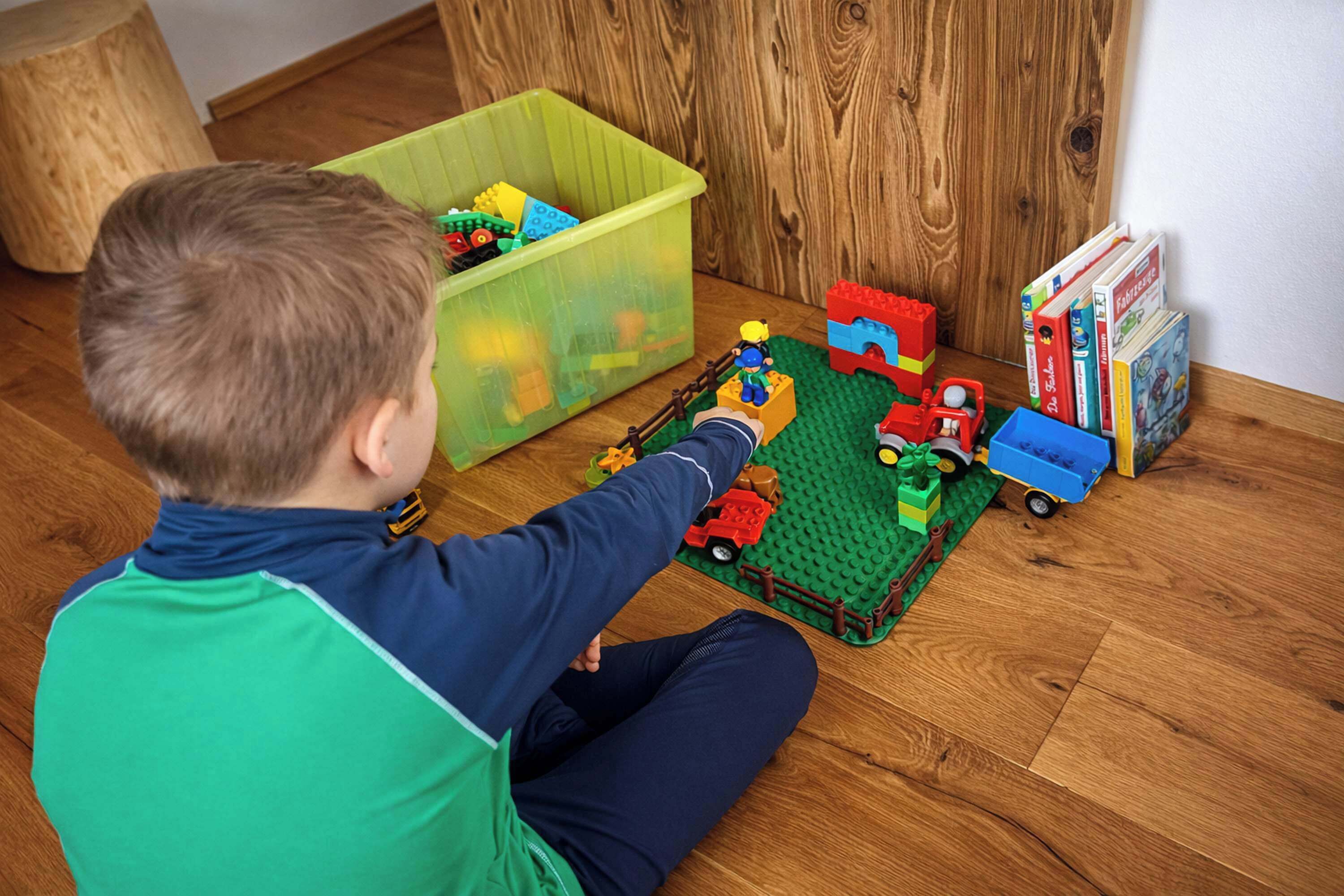 Ein Kind spielt mit LEGO auf einem Holzboden, in der Nähe steht ein grüner Mülleimer und Bücher.