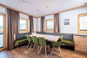 A room with a wooden dining table, green upholstered chairs, a bench, wooden cabinets, and large windows with curtains. - BEIM LOIS Isskogelblick – 2 Floors & Sauna Access (Gerlos)