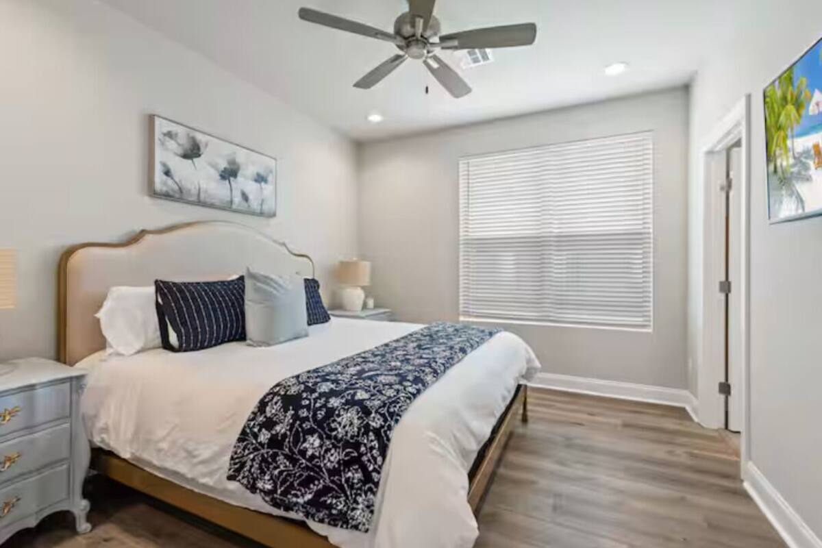 A bedroom with a bed, nightstands, a ceiling fan, a window with blinds, and a painting on the wall.