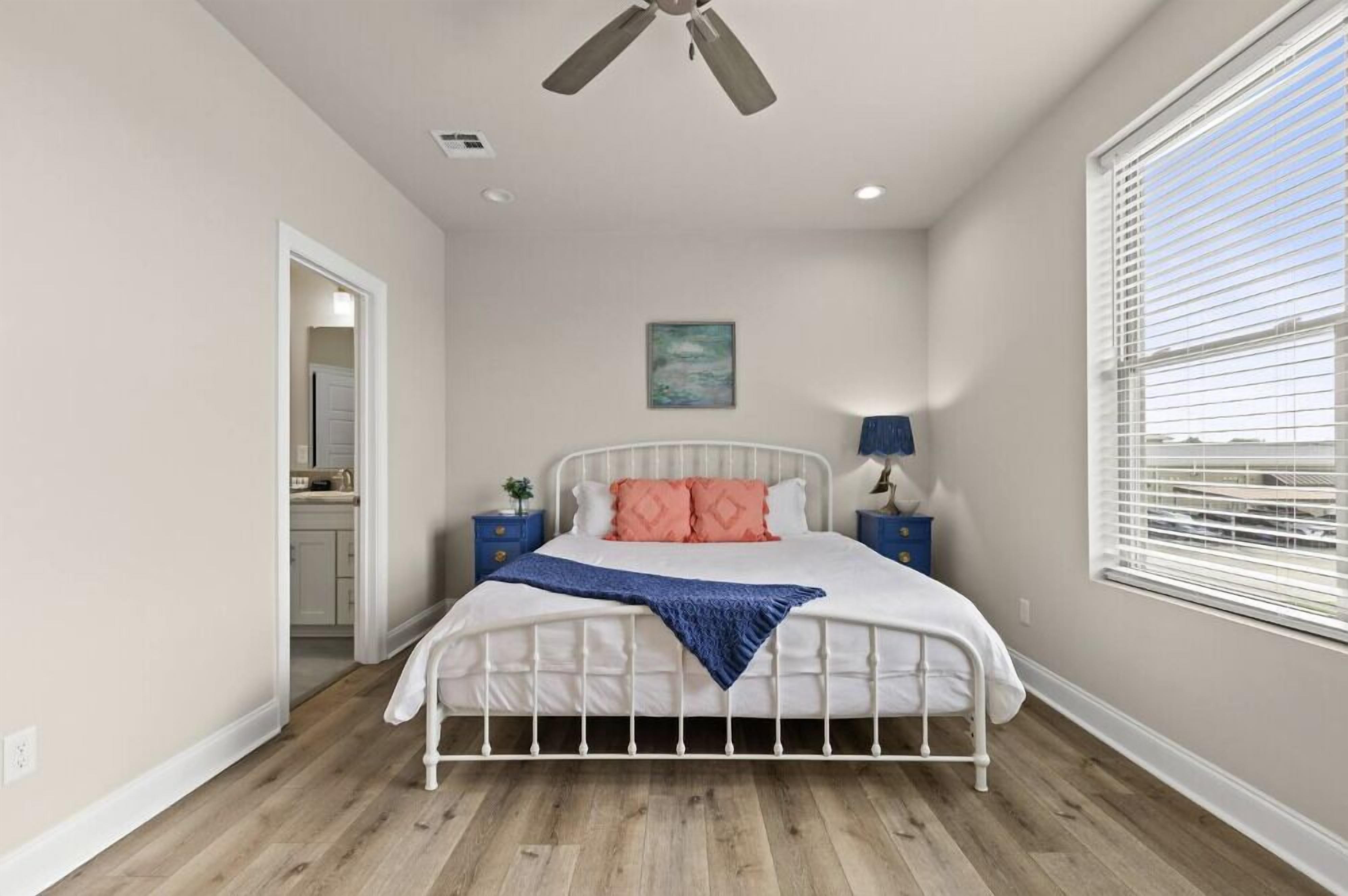 A bedroom with a bed, a ceiling fan, a window with blinds, a nightstand, and a painting on the wall.