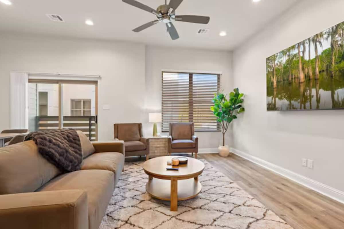 A living room with a sofa, chairs, a coffee table, a ceiling fan, and a large window with blinds.