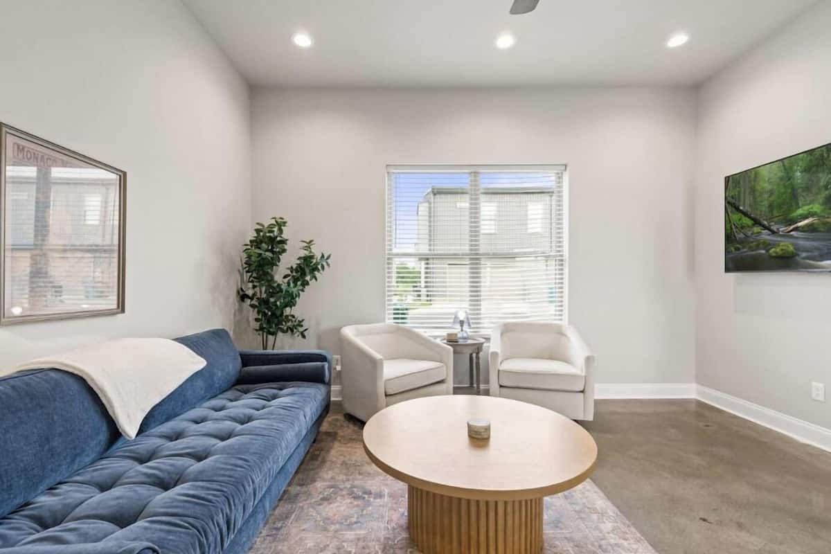 A living room with a blue sofa, two white chairs, a round coffee table, and a large window with blinds.