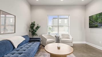 Un salon avec un canapé bleu, deux fauteuils blancs, une table basse ronde et une grande fenêtre avec des stores.