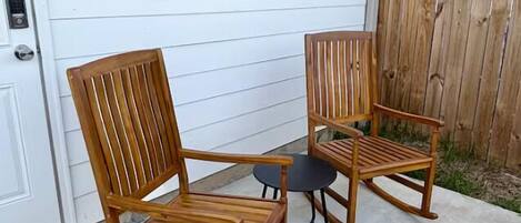 A wooden rocking chair and a small round table outside a house with white siding and a wooden fence.