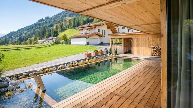 A wooden deck with a clear pool, a mountain view, and a house in the background. - RACING Suite – Terrace & Sauna Access (Kleinboden)
