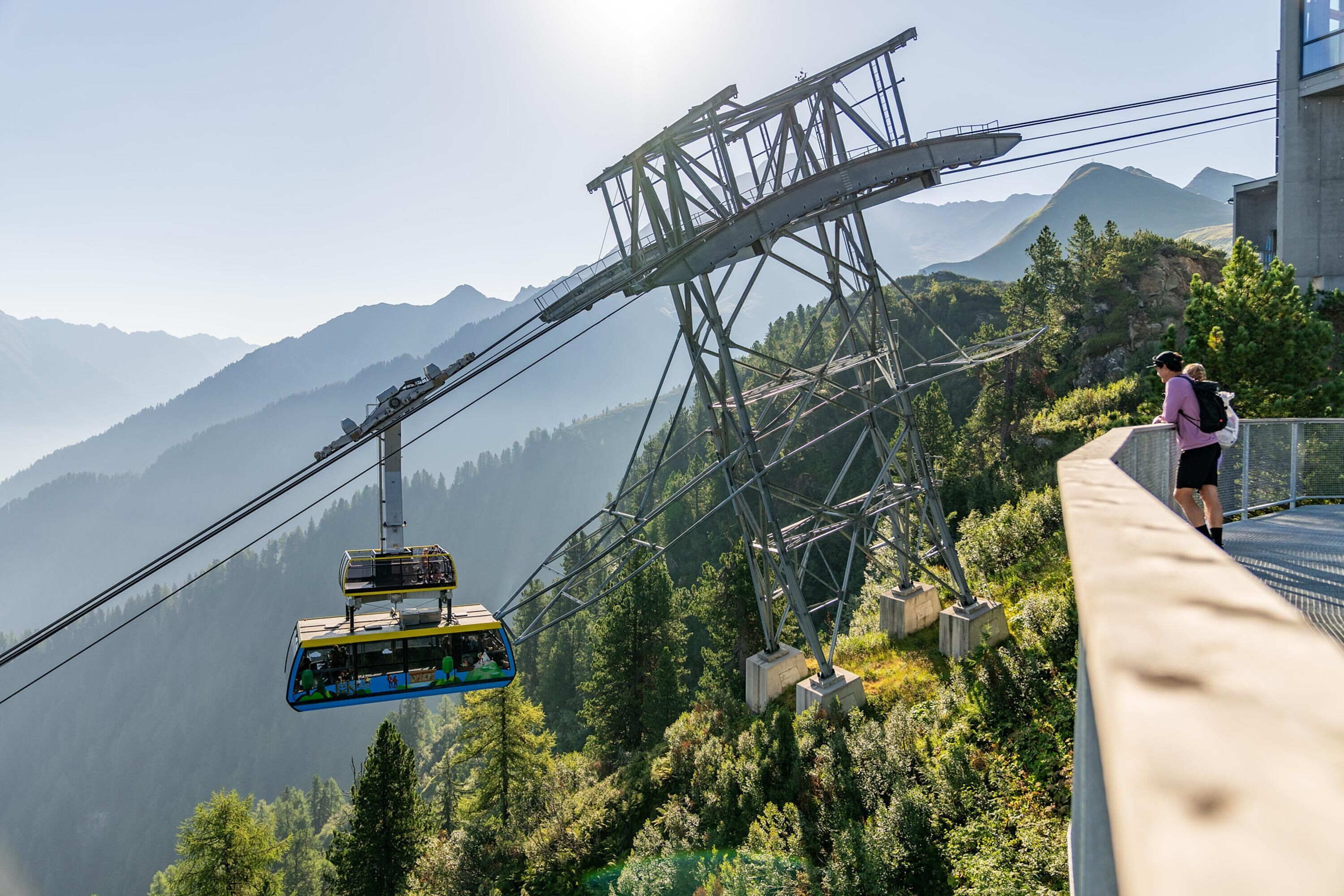 Un téléphérique avec une cabine pour passagers suspendue entre deux pylônes, une personne debout sur une plateforme et un paysage montagneux en arrière-plan.