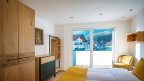 A bedroom with a bed, wooden wardrobe, a small wooden cabinet, a yellow chair, and a view of houses through a glass door. - ROFANLODGE Gutschlhofer – A house with a view, a terrace and parking (Mariatal)