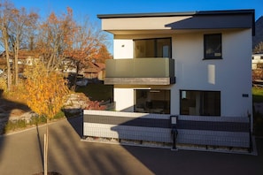 Une maison moderne à deux étages, dotée d’un balcon, d’une allée pavée et entourée d’un feuillage automnal.