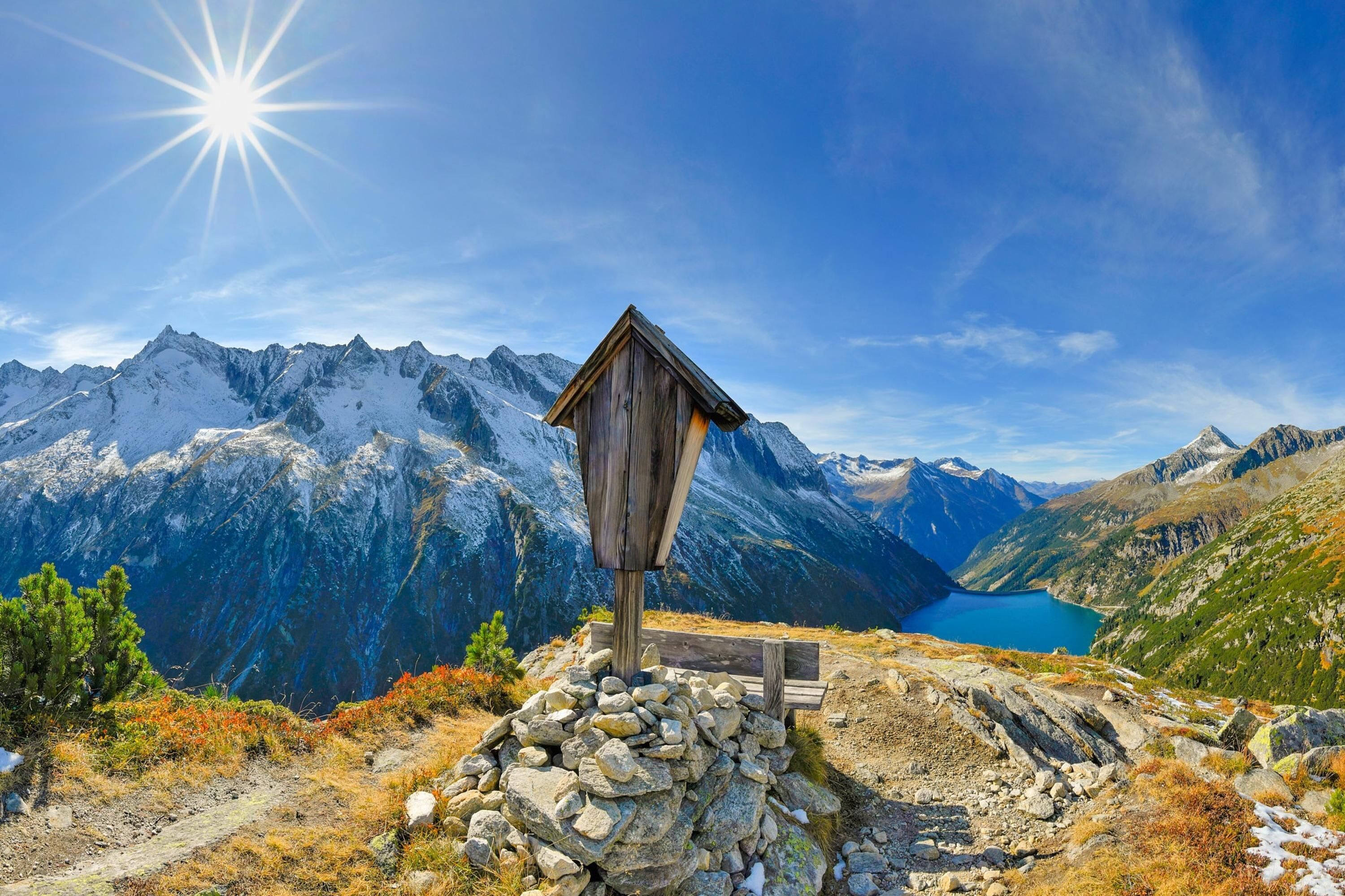A wooden mountain hut with a stone base, surrounded by rocky terrain and a lake, with snow-capped mountains in the background.