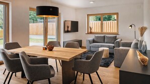 Ein modernes Wohnzimmer mit einem Holzespeisetisch, grau gepolsterten Stühlen, einem grauen Sofa, einer schwarzen Stehlampe und einem an der Wand montierten Fernseher.