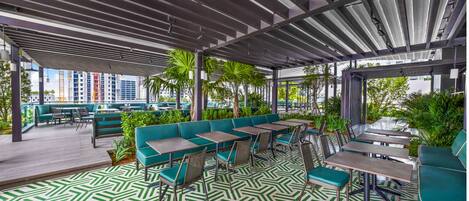 An outdoor seating area with green upholstered benches and tables, a patterned floor, and a view of buildings in the background.