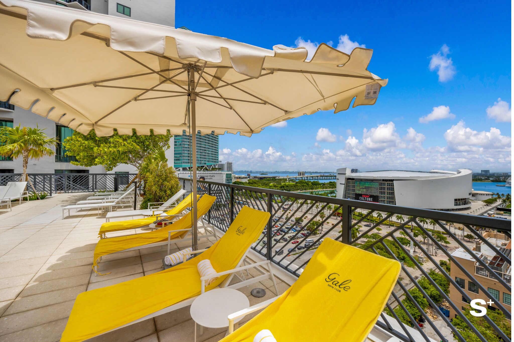 A rooftop lounge with yellow lounge chairs and a large umbrella, overlooking a cityscape with buildings and a body of water.