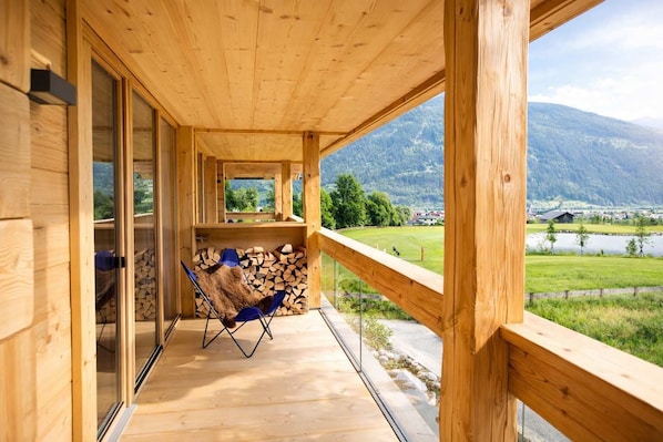 A wooden balcony with a view of a lake and mountains, a chair, and a stack of firewood. - MOUNTAIN Suite – Balcony & Sauna (Kleinboden)