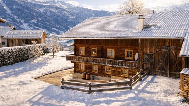 Un chalet in legno con un balcone, un muro in pietra e un paesaggio innevato con montagne sullo sfondo.