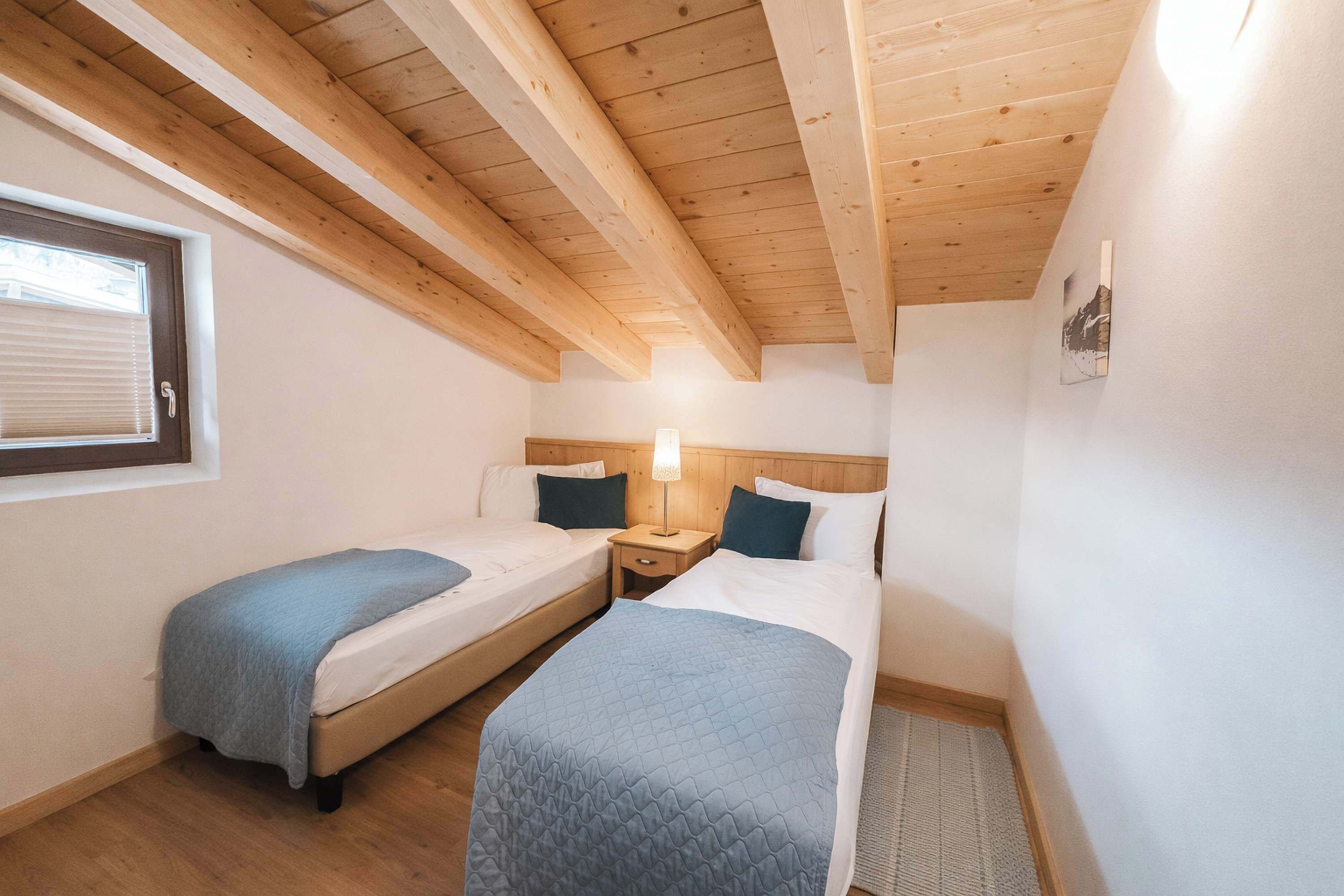 Une chambre avec deux lits, des poutres en bois au plafond, une fenêtre avec des stores et une lampe de chevet.