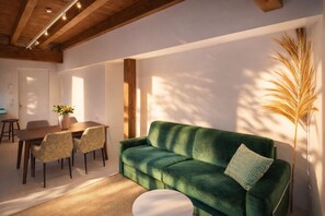 A room with a green sofa, a dining table with chairs, a white round table, and a tall plant.