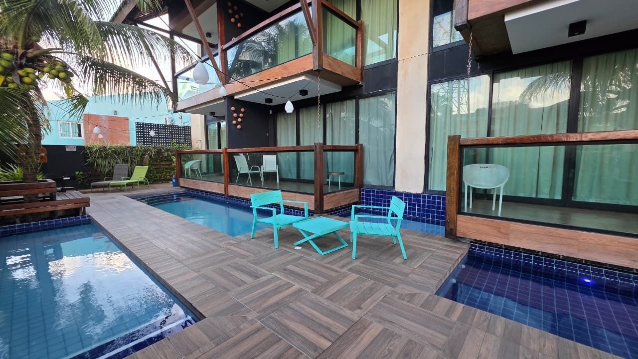 Un espace piscine extérieur moderne, doté d’une terrasse en bois, d’un coin salon avec des chaises et d’un bâtiment à deux étages avec de grandes fenêtres.