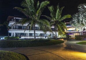 A modern hotel with a pool, palm trees, and a well-lit entrance.