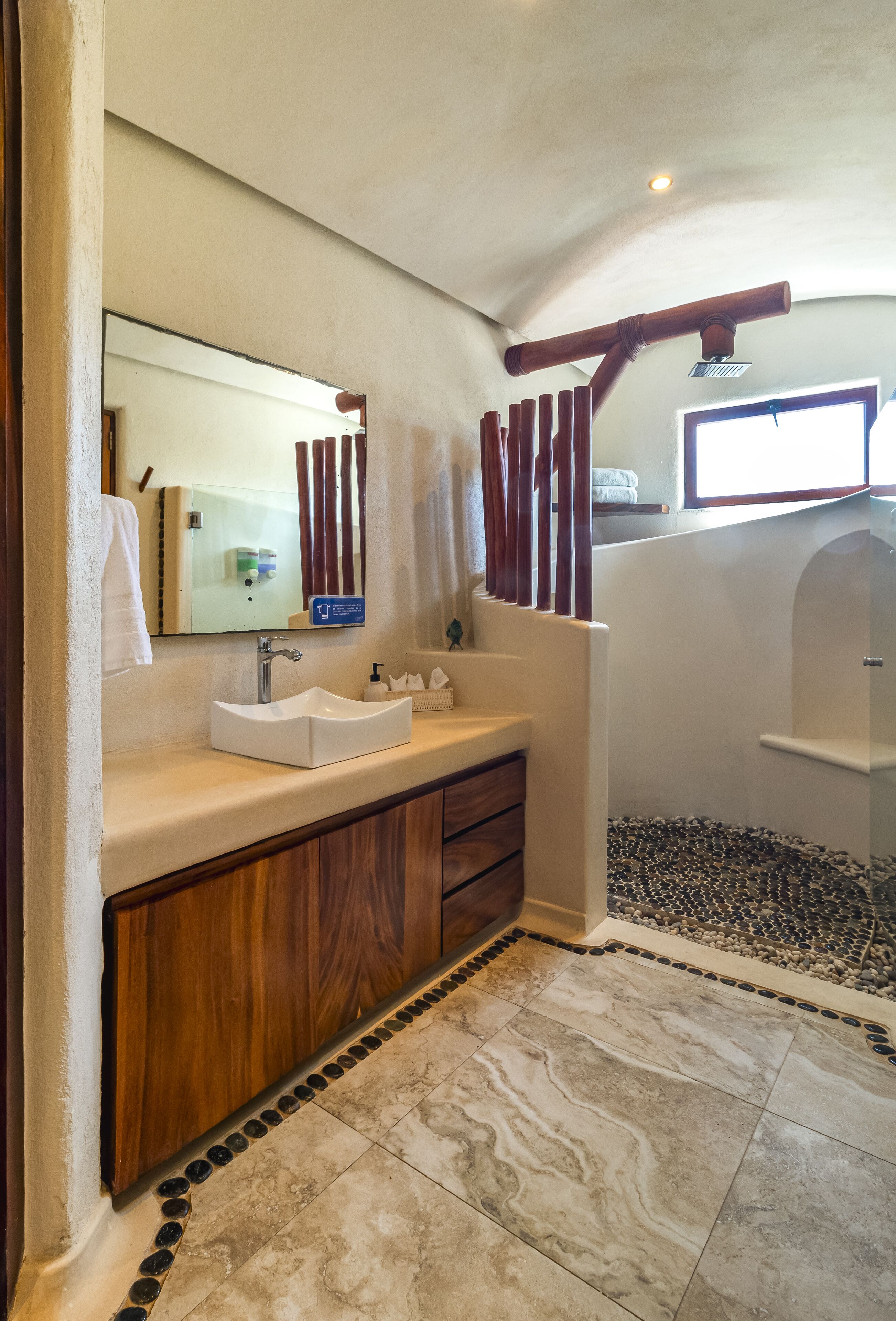 Baño con lavamanos blanco, tocador de madera, espejo y zona de ducha.