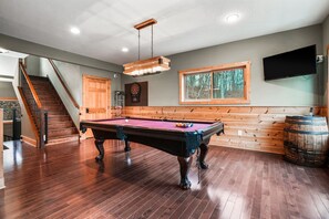 Une table de billard dans une pièce dont le sol est en bois, avec un banc en bois, une fenêtre donnant sur des arbres et un escalier.