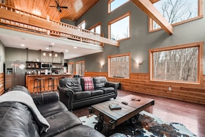A spacious living area with a high ceiling, a kitchen, and large windows offering views of the outdoors. - Starr & Sky Lodge | Hocking Hills, Hot Tub, Sauna (Logan)