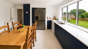 A modern kitchen with a dining table, chairs, a refrigerator, and a large window offering a view of the outdoors. - Spacious Cotswolds Home-Large Garden-Games Room (Finstock)