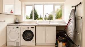 A compact kitchen with a washing machine, a sink, and a window overlooking greenery. - Spacious Cotswolds Home-Large Garden-Games Room (Finstock)