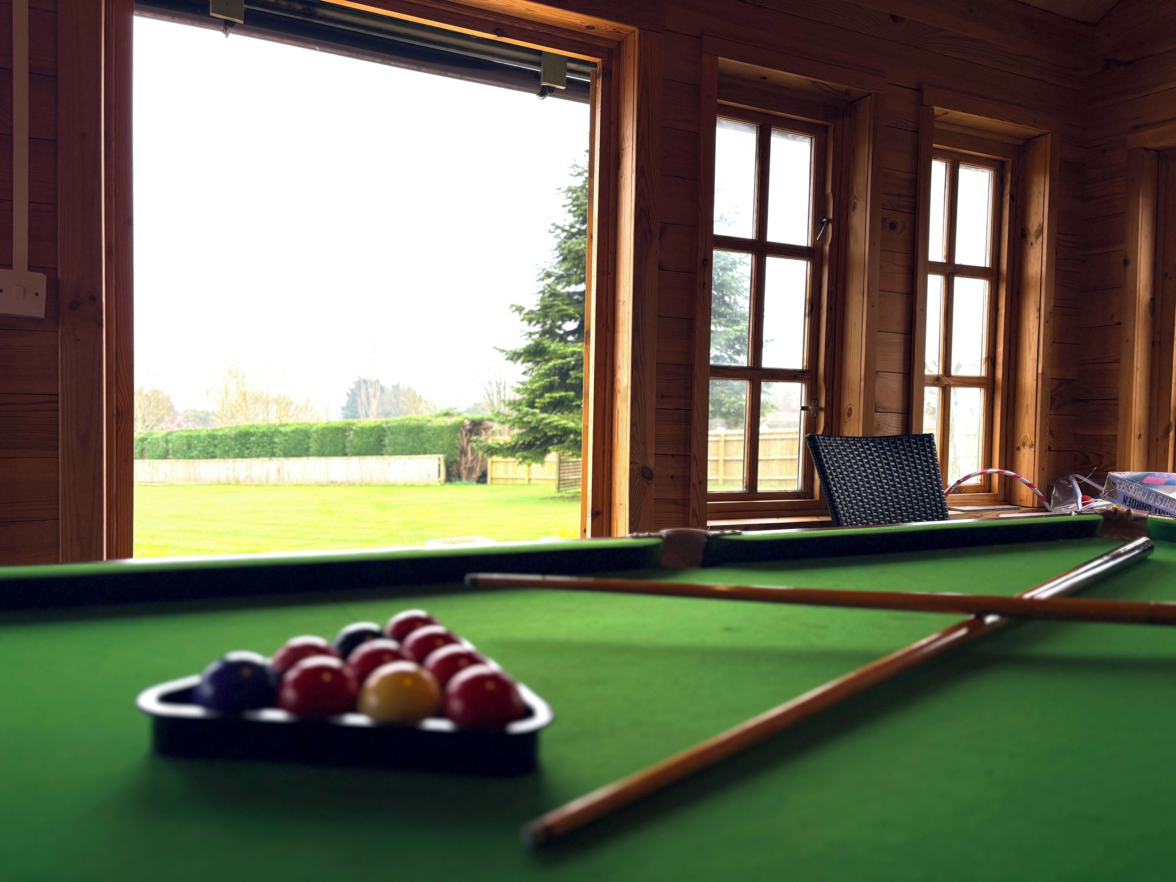 A pool table with balls, a cue stick, and a laptop in a room with wooden walls and large windows.