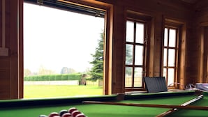 Une table de billard avec des billes, une queue et un ordinateur portable, dans une pièce aux murs en bois et aux grandes fenêtres.