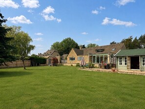 A residential area with a well-maintained lawn, a covered patio area, and houses with pitched roofs. - Spacious Cotswolds Home-Large Garden-Games Room (Finstock)