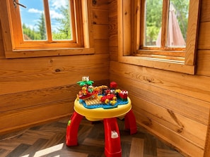 Interior de uma cabana de madeira com um espaço infantil, incluindo uma mesa LEGO colorida e um pequeno banquinho.