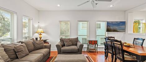 A living room with a sofa, armchair, ottoman, dining table with chairs, and a ceiling fan.