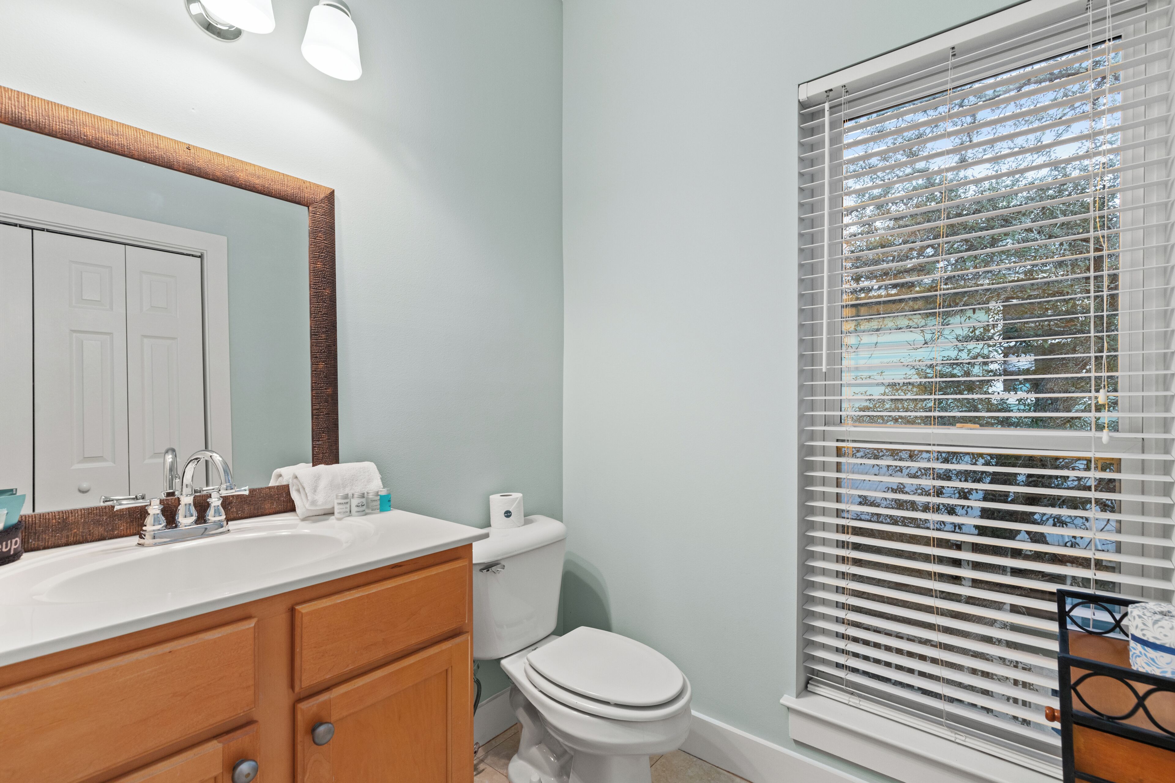Une salle de bain équipée d’un lavabo, d’un miroir, de toilettes et d’une fenêtre avec des stores.