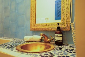 Un baño con suelo de baldosas estampadas, lavamanos con grifo dorado, una botella de jabón de manos y un espejo con marco decorativo.