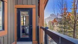 A modern building with a black door, a balcony with a wooden railing, and a view of a mountain and other buildings in the background. - Chic Rocky Mountain 2BR w/ Hot tub and Pool (Canmore)