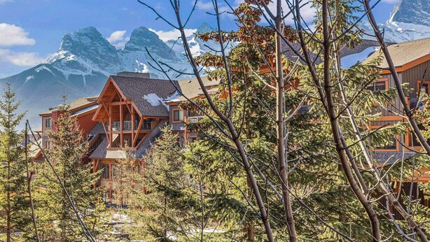 A lodge with a prominent mountain backdrop, wooden architecture, and a view of the surrounding forest. - Chic Rocky Mountain 2BR w/ Hot tub and Pool (Canmore)