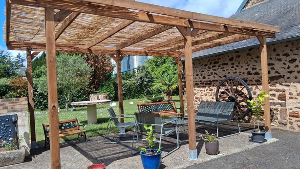Een houten pergola met zitplaatsen, plantenpotten en een stenen muur met een wiel.