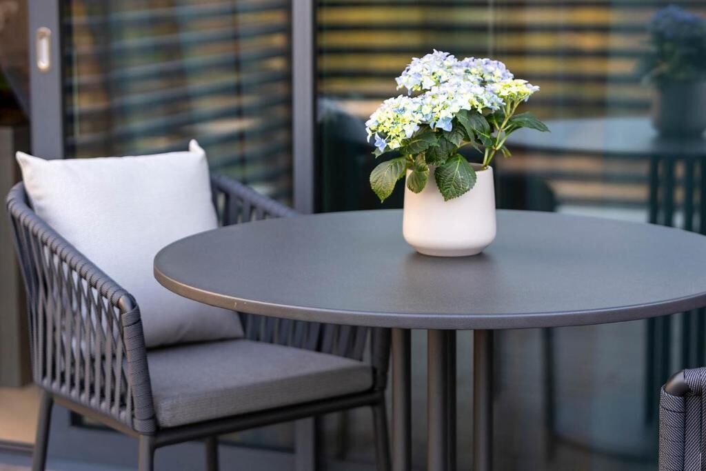 Een ronde tafel met een witte vaas met bloemen, een grijze stoel met kussentje en een glazen deur op de achtergrond.