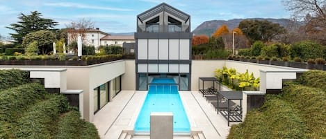 A modern pool area with a geometric pool, surrounded by neatly trimmed hedges and a clear sky.