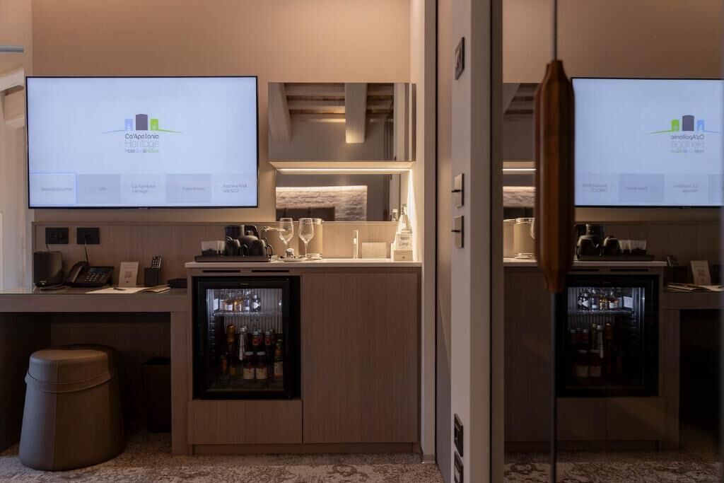 Habitación de hotel con minibar, escritorio con computadora y dos televisores mostrando el logotipo del hotel.