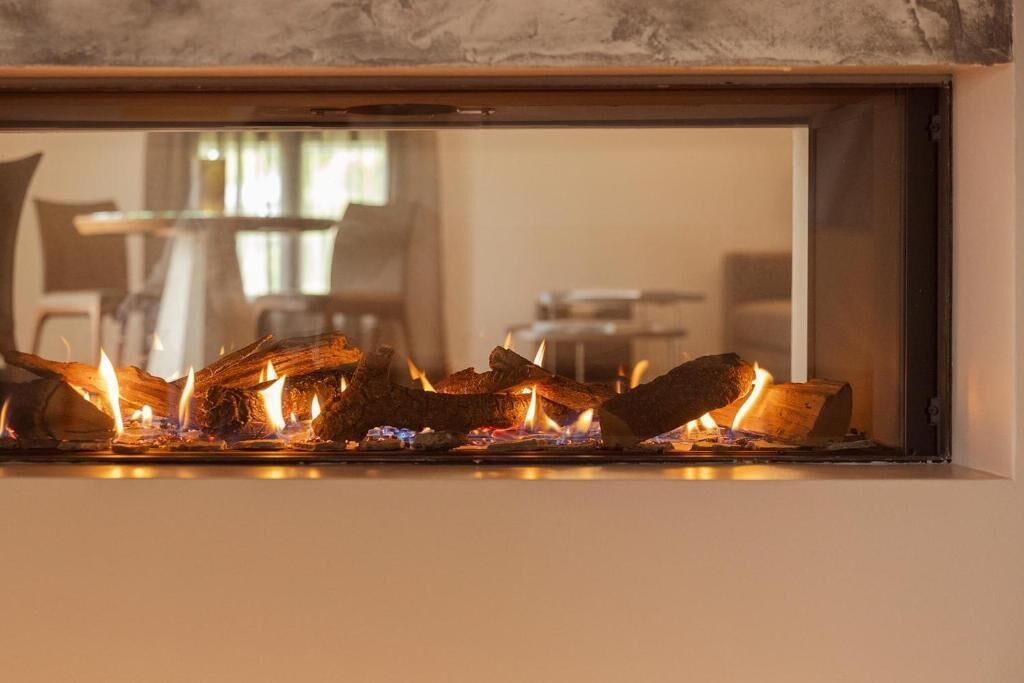 A fireplace with burning logs, a dining area with chairs and a table, and a window with a view of the outdoors.
