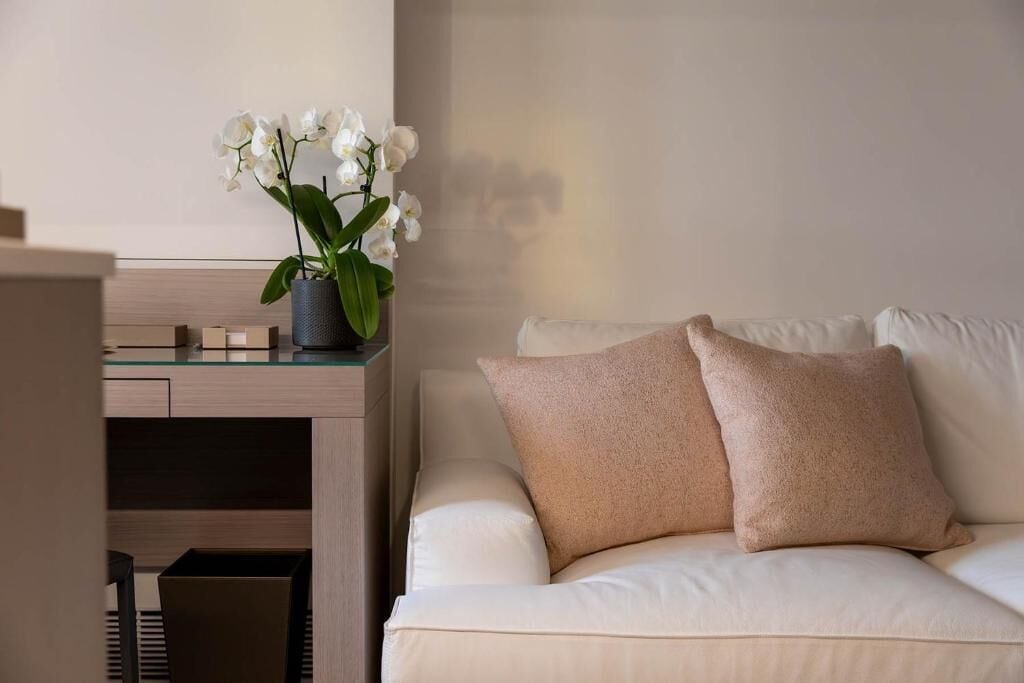 Una sala de estar moderna con un sofá blanco, dos cojines color beige, una mesa de centro de vidrio y una orquídea blanca en maceta.