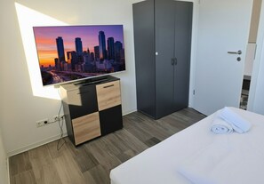 A hotel room with a bed, a TV mounted on the wall, a wardrobe, and a view of a cityscape. - Free Parking & Balcony upper floor flat - APOLONIAliving (Karlsruhe)