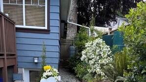 Eine Terrasse mit einem runden Tisch und zwei Stühlen, umgeben von einem Garten mit verschiedenen Pflanzen, mit einem blauen Haus im Hintergrund.