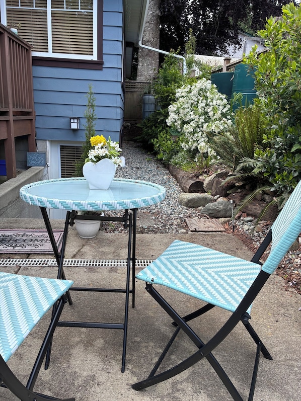 A patio with a round table and two chairs, surrounded by a garden with various plants and a blue house in the background.